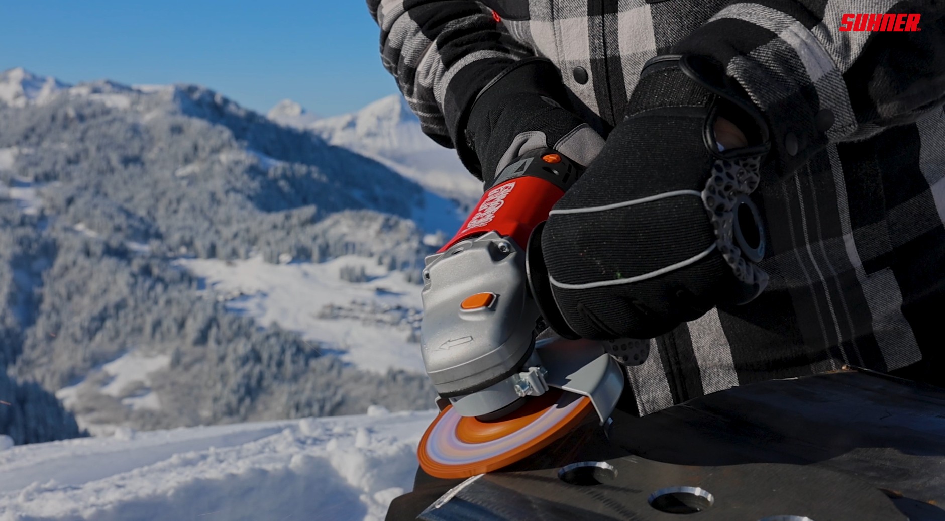 Close-up of a snowboarder’s gloved hands gripping an orange-gray power tool mounted to a snowboard, with snowy mountains in the background and a red SUUNNER logo in the corner.